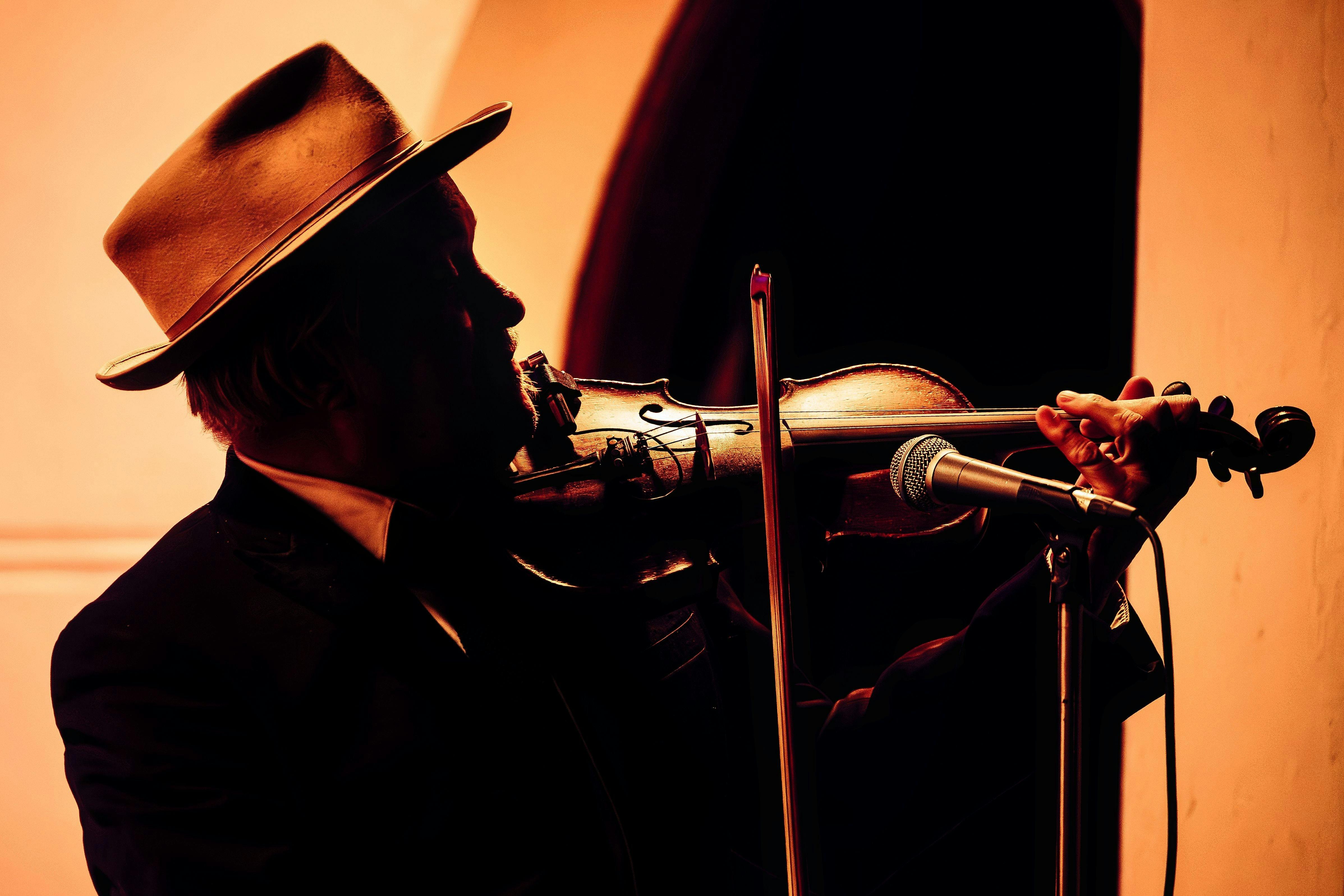 A man in a fedora is silhouetted in profile playing a violin. The lighting is orange and yellow