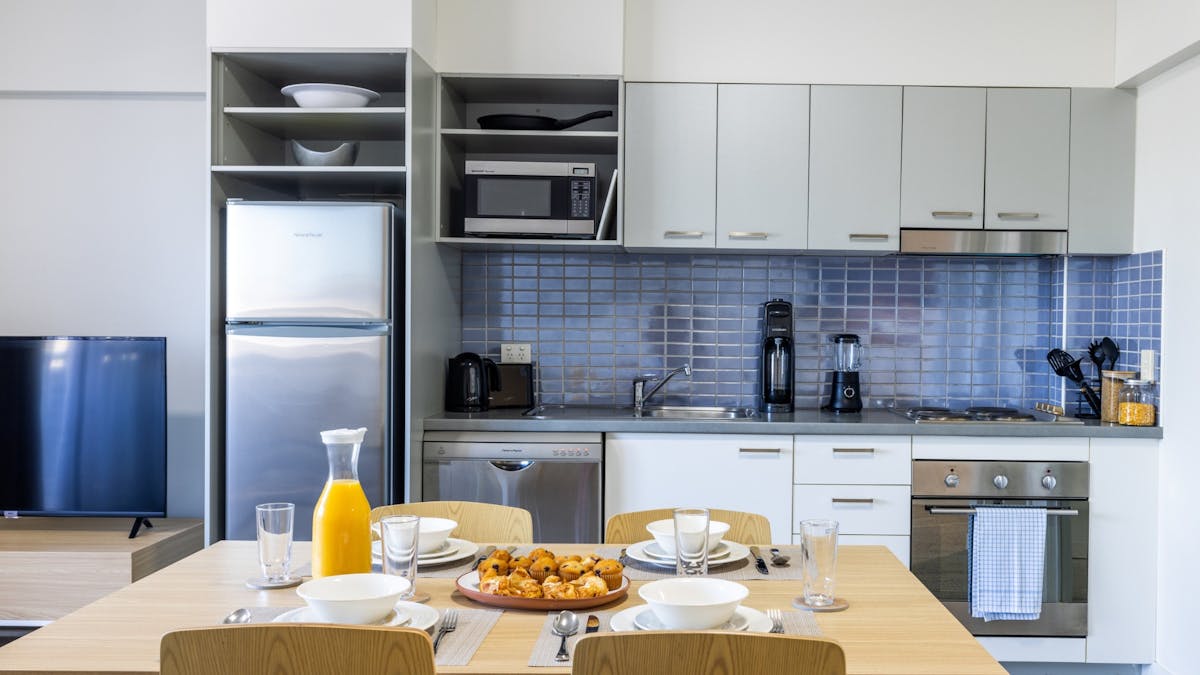 Kitchen cupboards, blue splashback tiles, a full sized silver fridge and a set dining table.