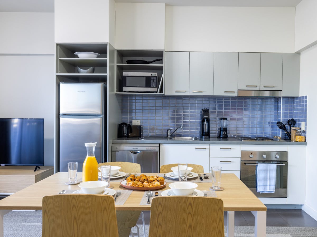 Kitchen cupboards, blue splashback tiles, a full sized silver fridge and a set dining table.