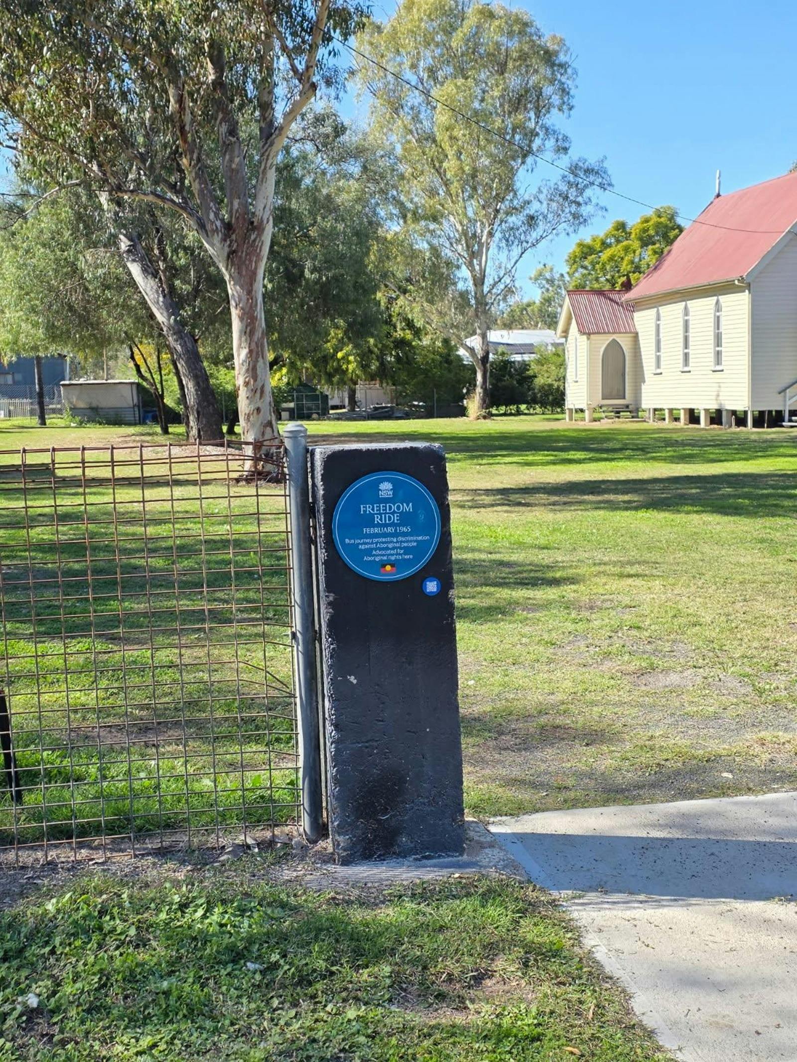 Blue plaque on fence post, in back ground is a church and park  area with trees