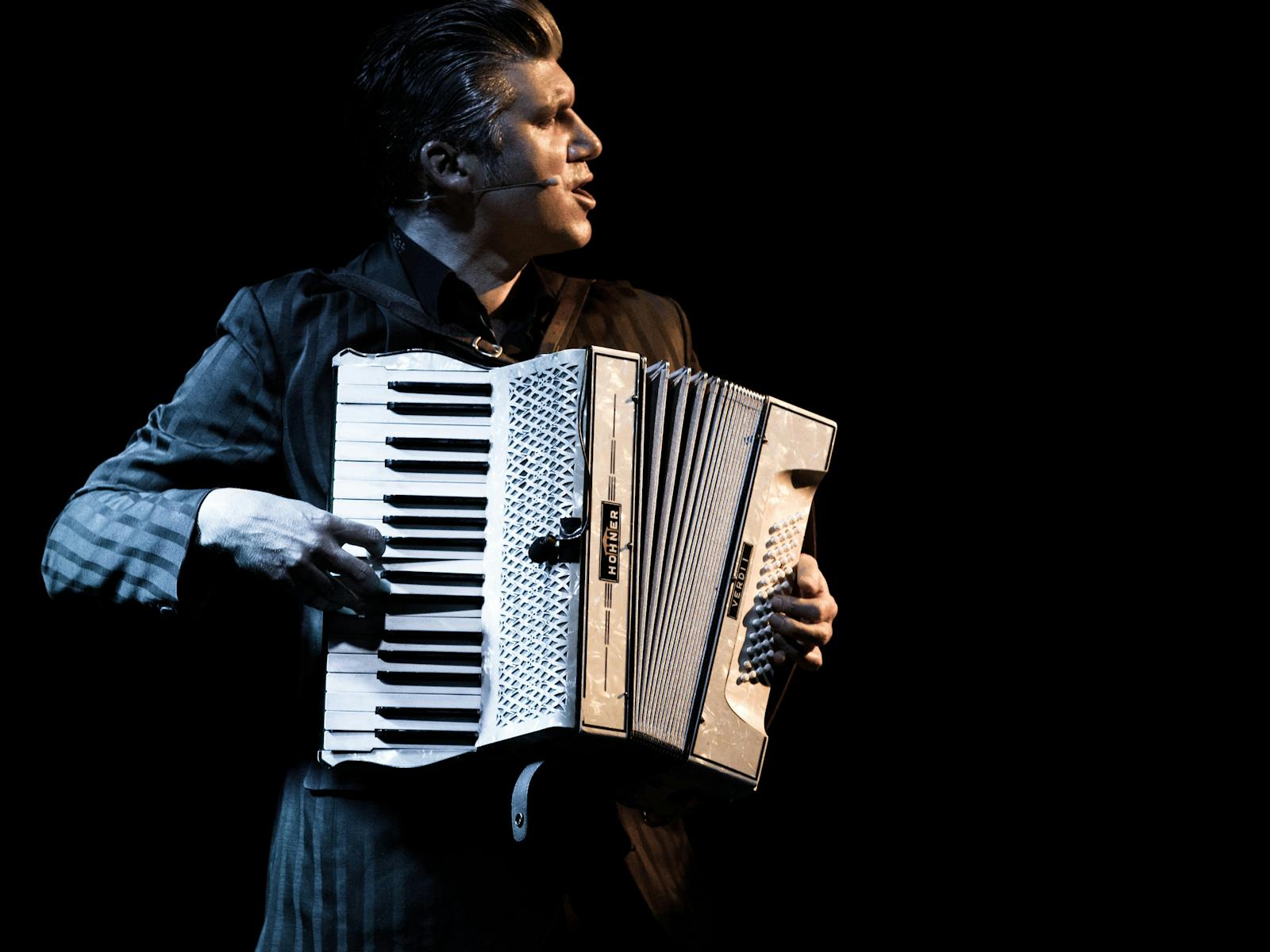 A man plays an accordion, bathed in a golden light and darkness.