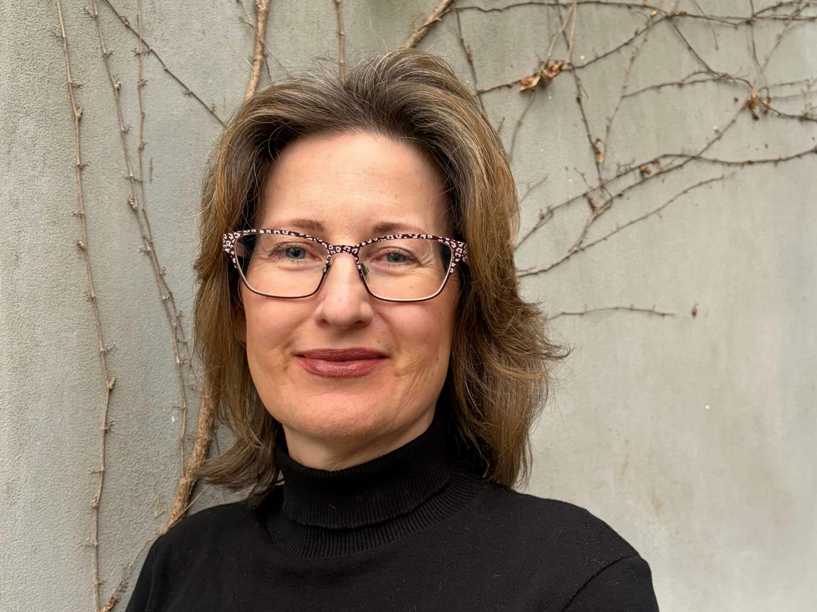 A woman with brown, wavy hair and glasses, wearing a black turtleneck.