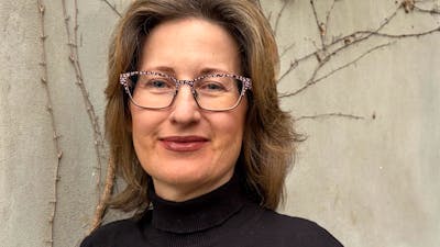 A woman with brown, wavy hair and glasses, wearing a black turtleneck.