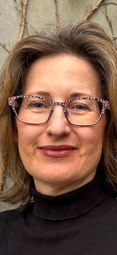 A woman with brown, wavy hair and glasses, wearing a black turtleneck.