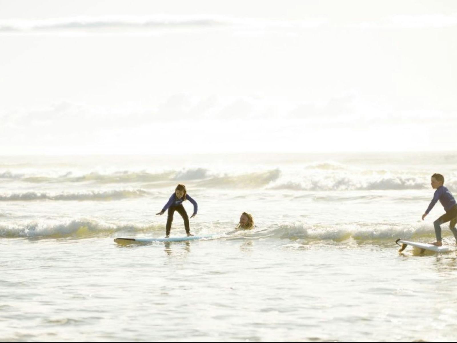 two kids standing up on boards on small wave with instructors in backgroundd