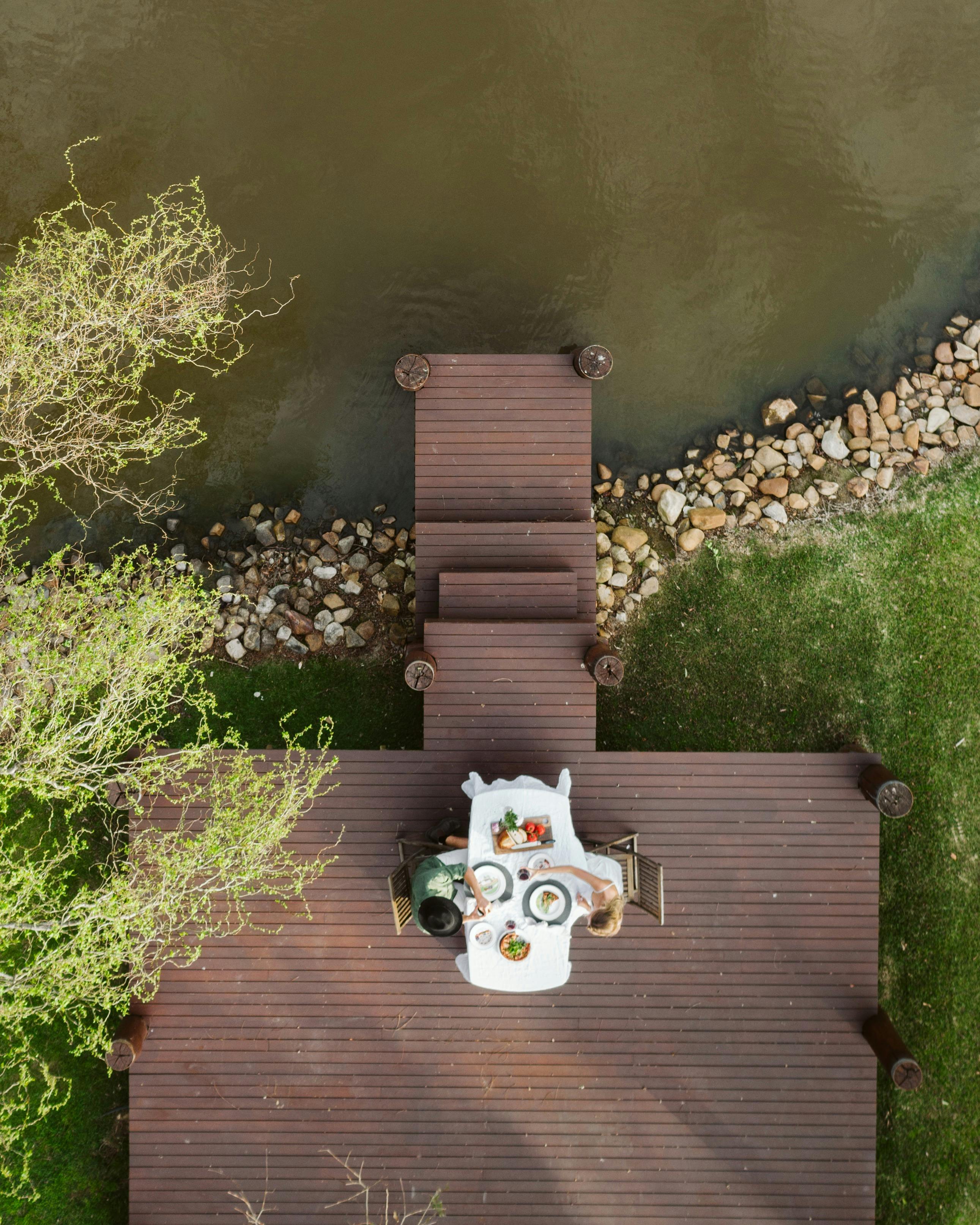 Image of airial view of deck that spans onto the lake. Couple enjoying a meal lakeside tree to left