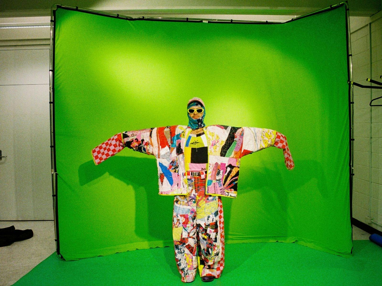 Man in large colourful suit with arms outstretched and standing in front of a green  screen