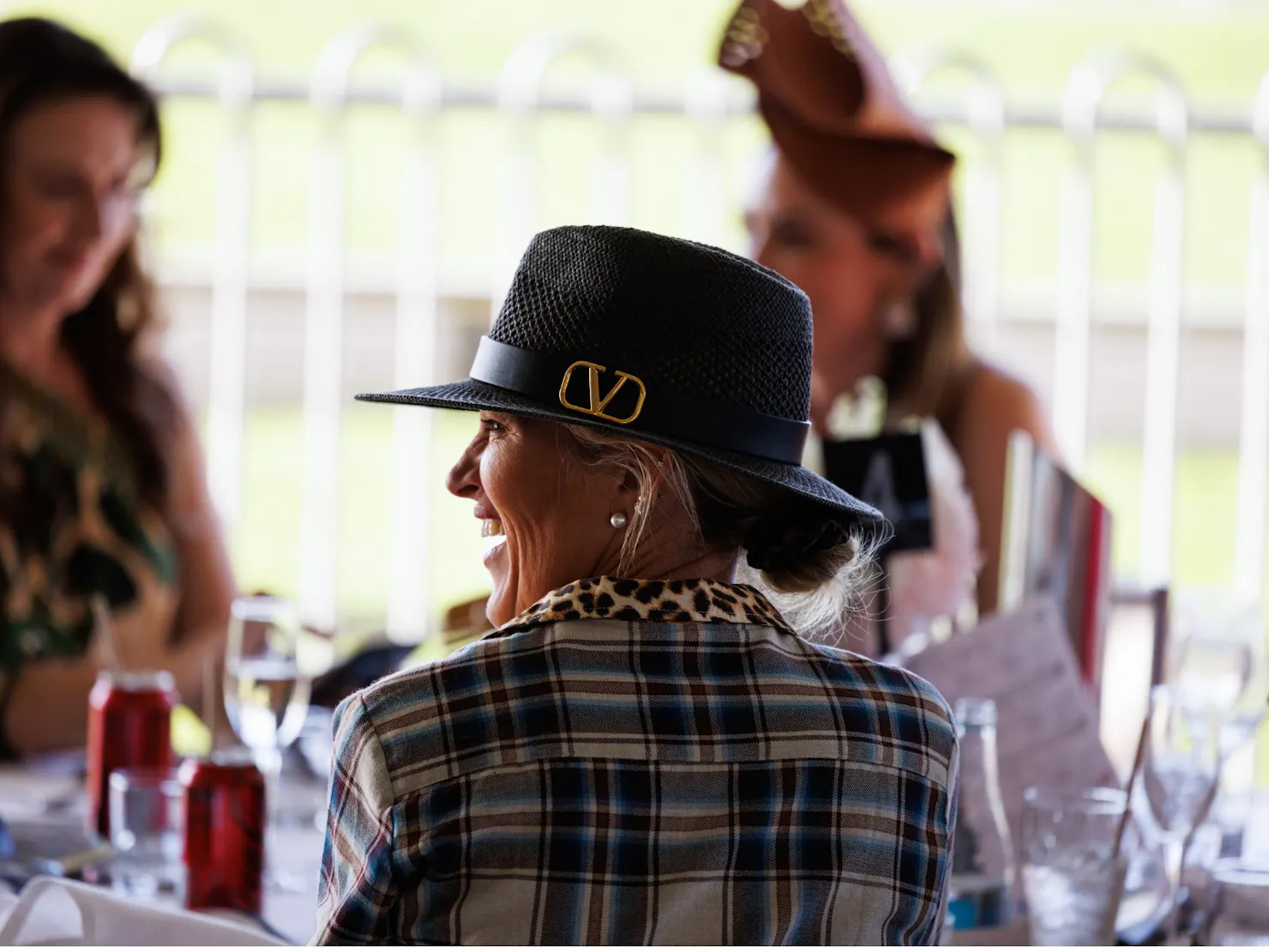 Side profile of a woman laughing, wearing a black hat with a gold buckle detail and a plaid jacket,