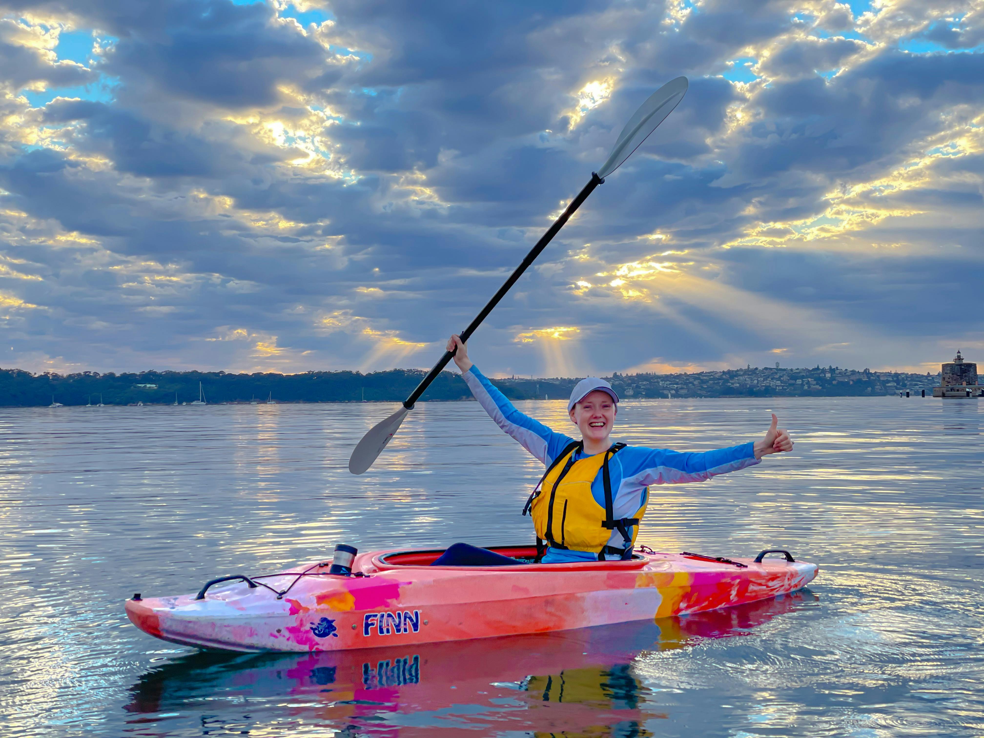 Laura Stone giving thumbs up from kayak with sun creeping through clouds on Sydney Harbour.