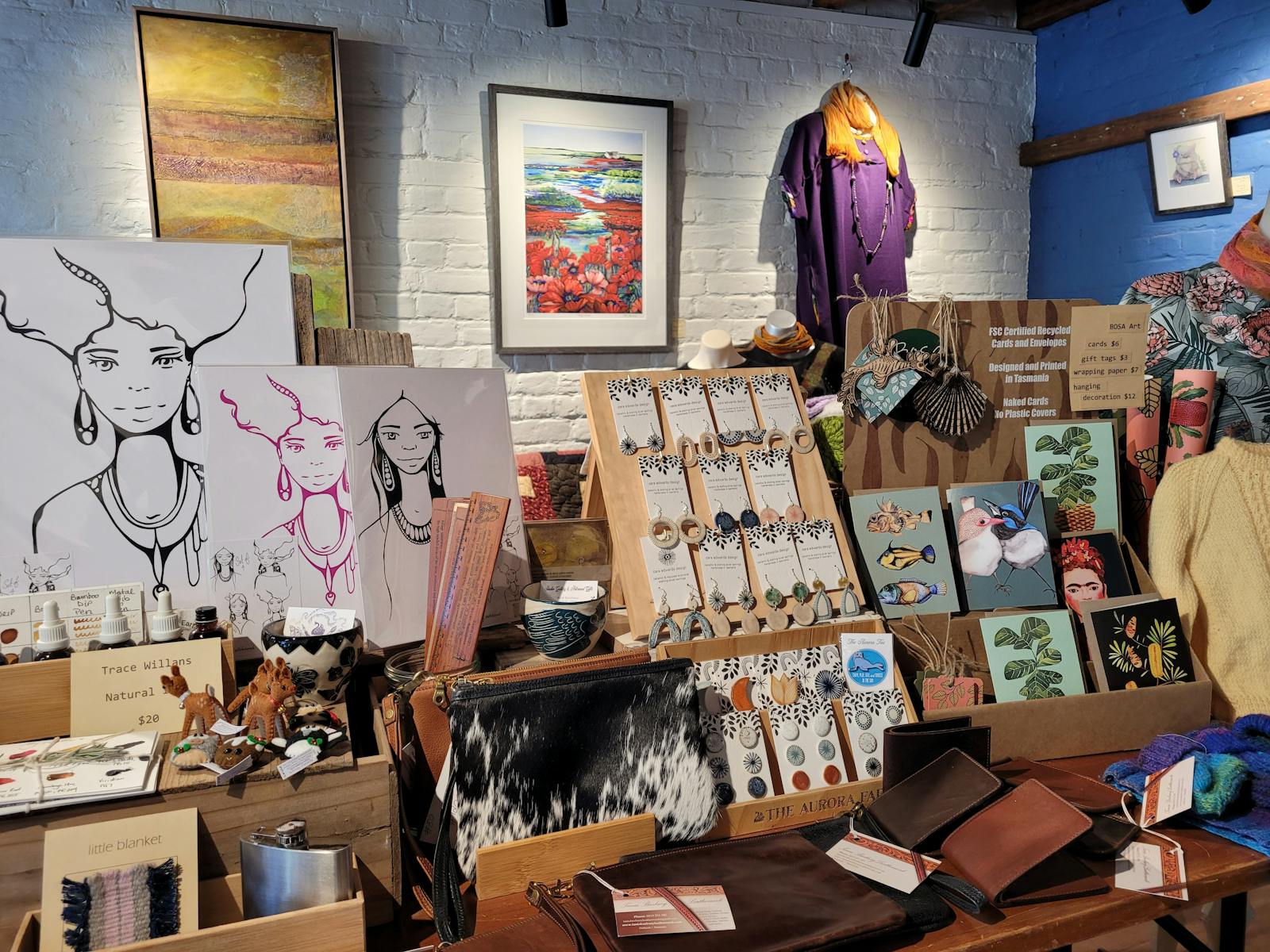 Art on white and blue walls, prints, earrings, bags and cards on display for sale on a central table