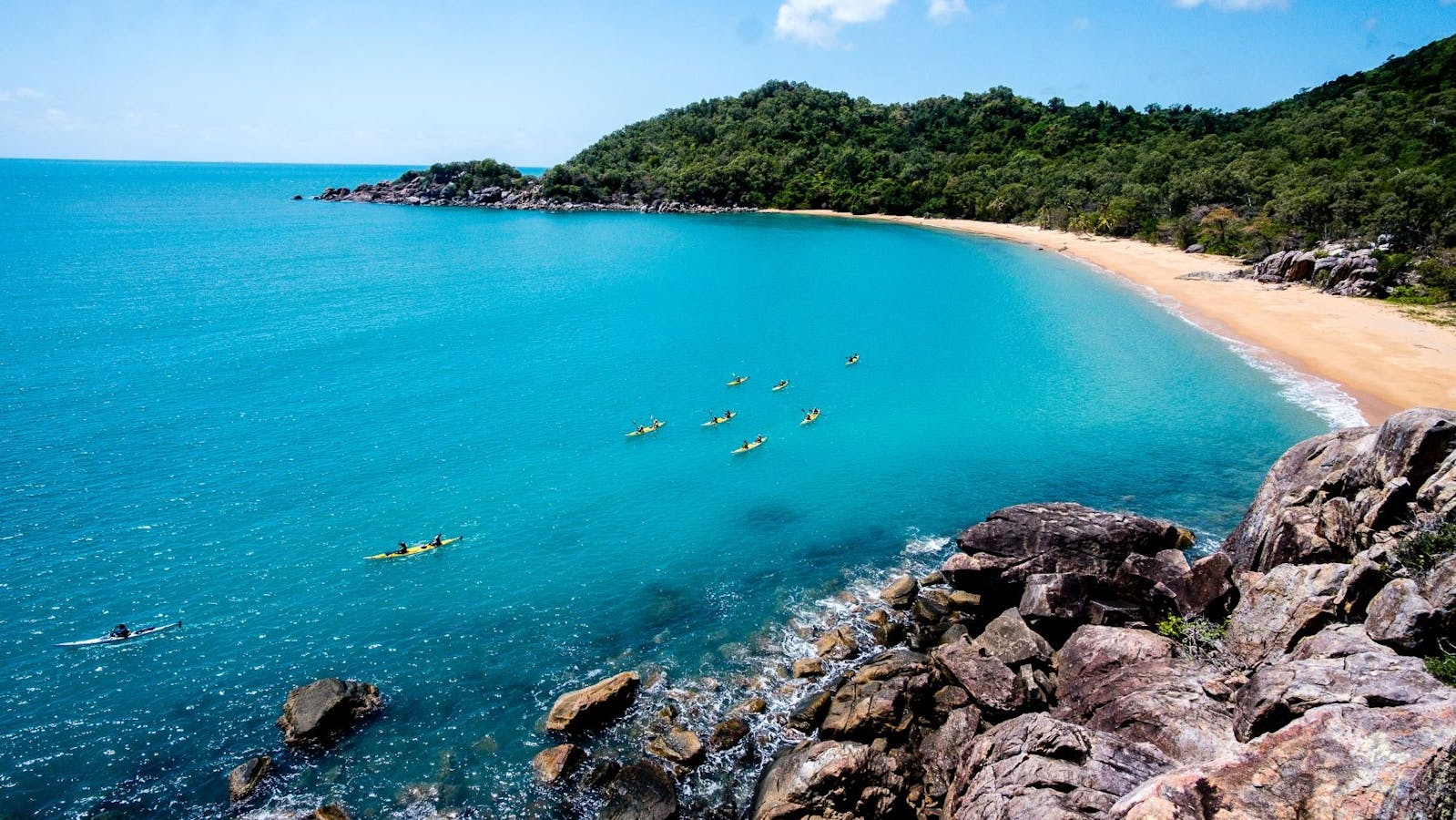 Sea Kayakers paddle across bright, clear blue water in front of a small bay flanked by rainforest Sea Kayakers paddle across bright, clear blue water in front of a small bay flanked by rainforest