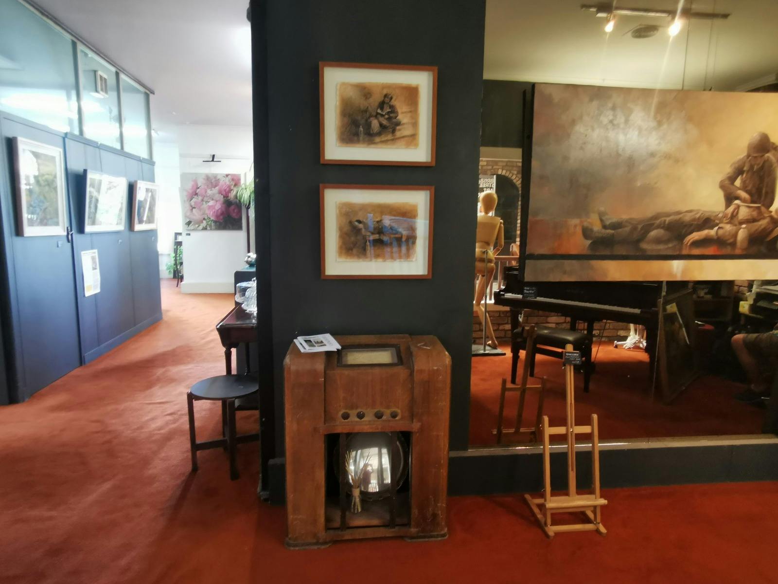 Interior view of an art gallery with framed realist artworks on dark walls, warm lighting and a heri