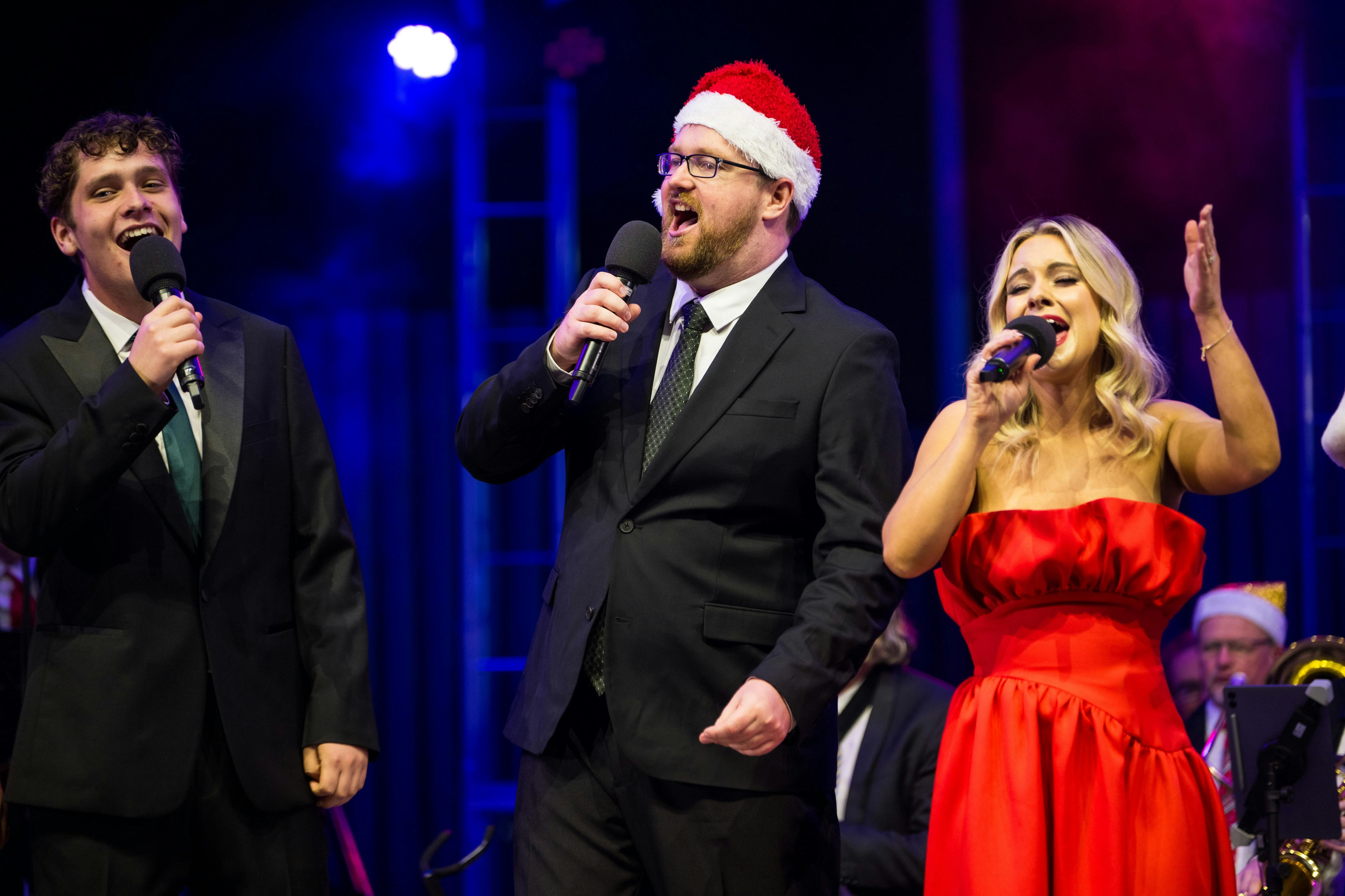 Man in santa hat and suit next to a woman in a red dress singing Christmas Carols on stage