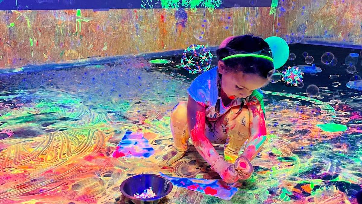 A little girl sat on the floor paying with glowing paint and bubbles at Messy Glow Co UV Paint Room