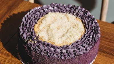 A round ube cake with purple frosting and a light shredded topping, placed on a wooden surface.