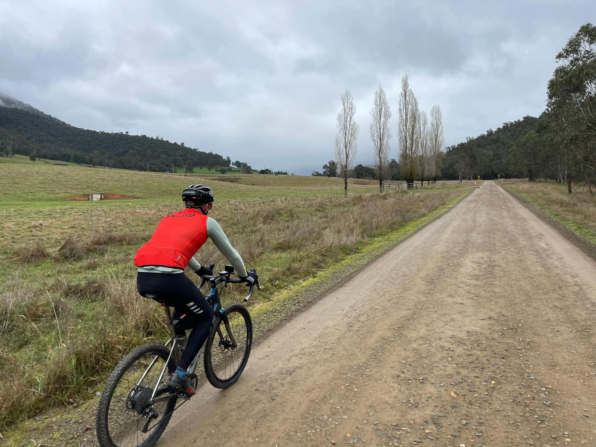 Cyclist with red vest on gravel road, farming country with poplar trees, hills with trees, grey sky