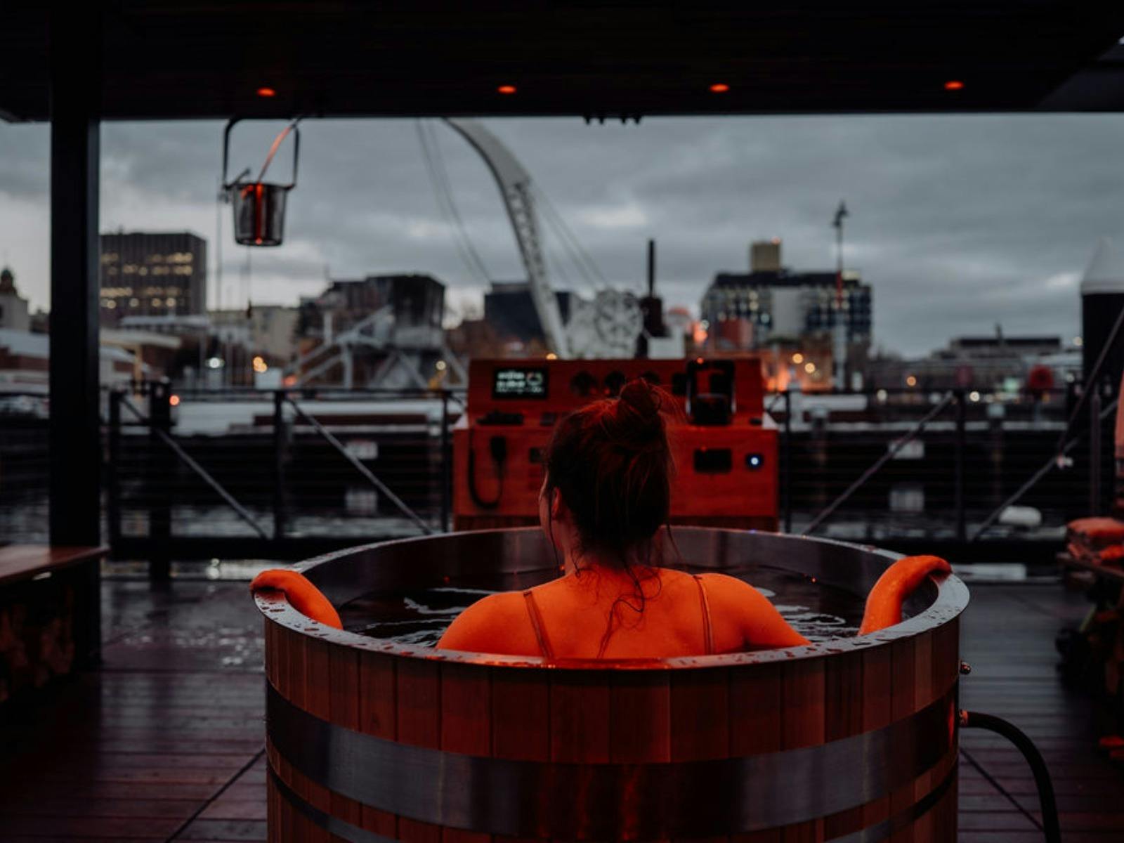 A woman in a wooden hot tub looks out at a city harbor and cranes at twilight.