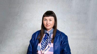 A person wearing a scarf, striped shirt, and blue leather jacket stands against a plain grey wall.