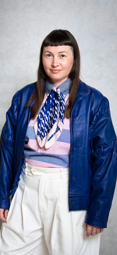 A person wearing a scarf, striped shirt, and blue leather jacket stands against a plain grey wall.