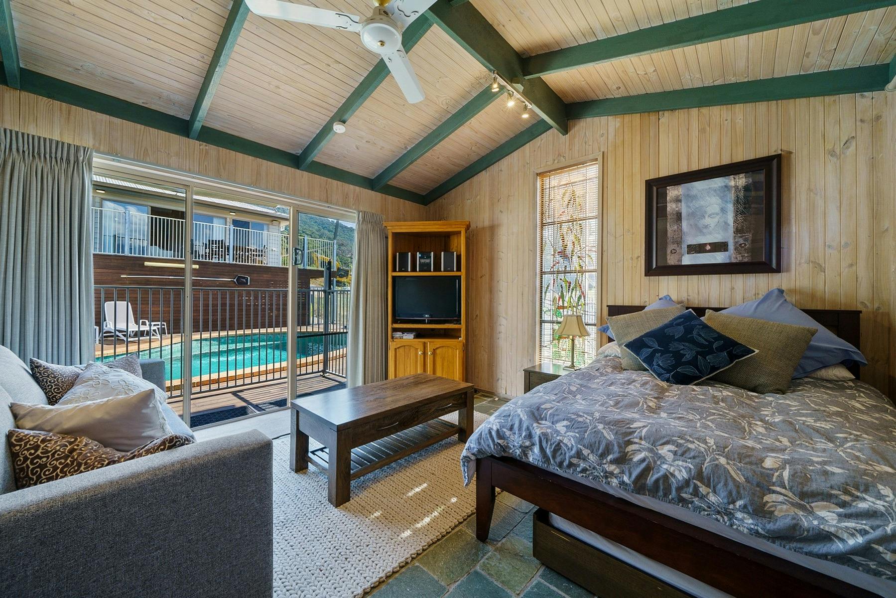 A pool house with a bed, couch and table. Swimming pool in the background outside.
