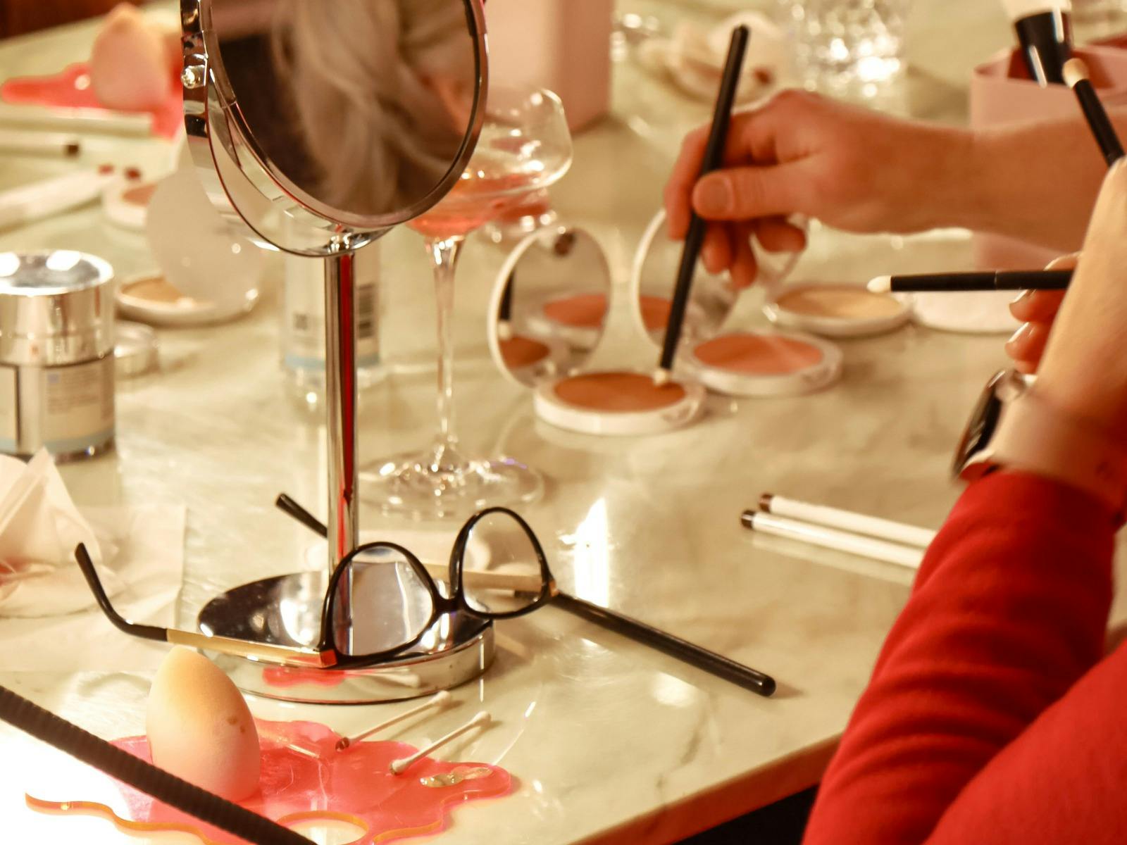 Hands applying blush with brushes at a beauty table with makeup, mirror, glasses and wine glass.