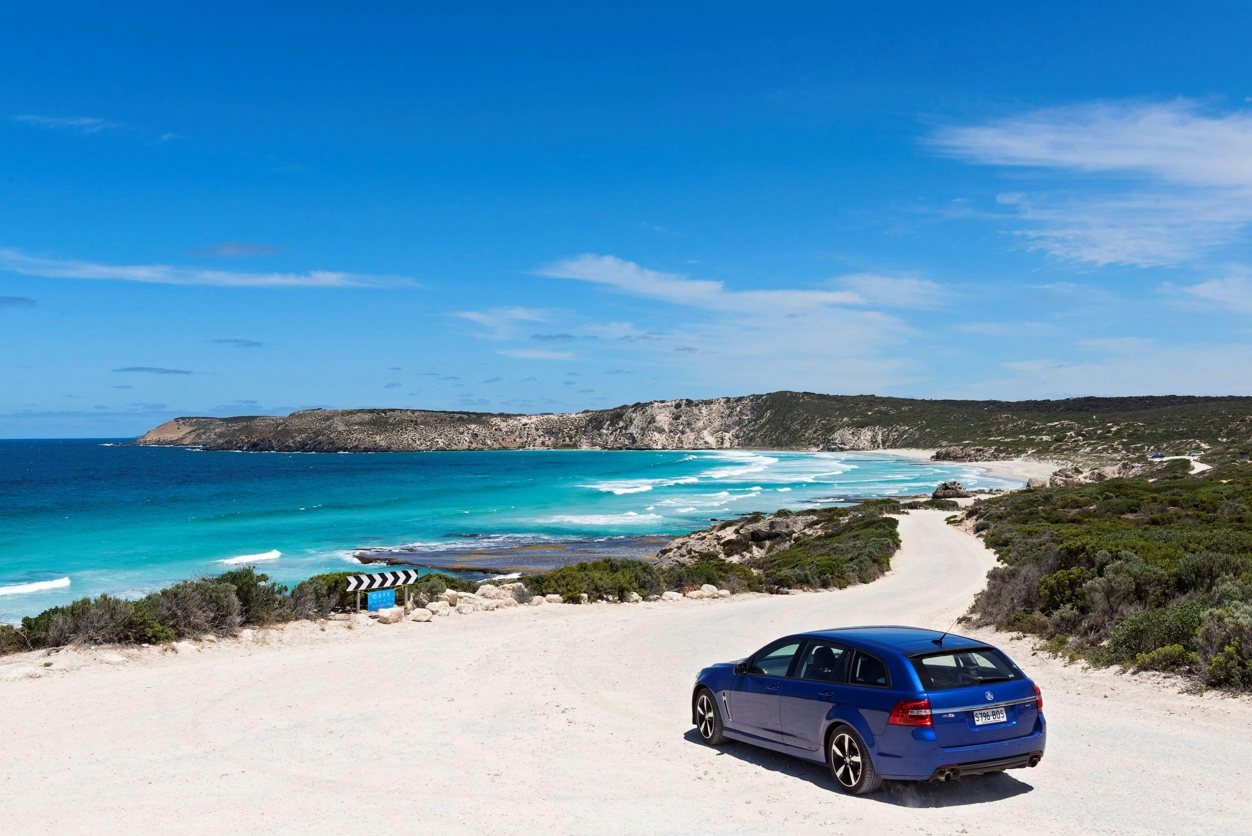 blue car driving down dirt road towards beautiful beach