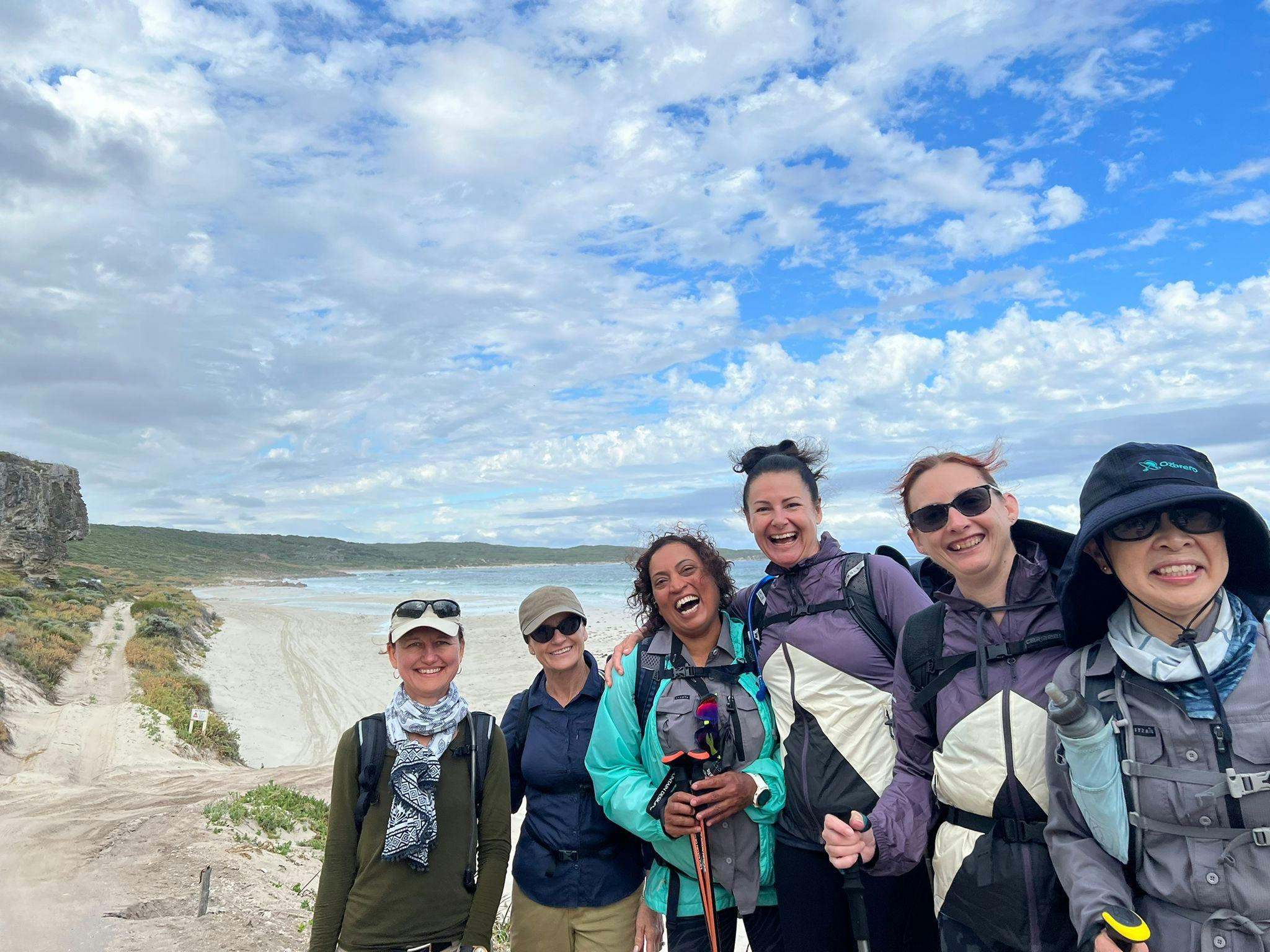 6 women smiling widely with a pristine beach behind them.