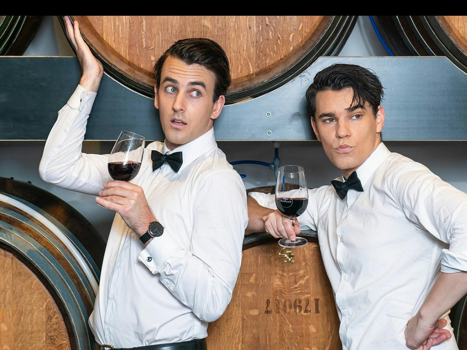 Two men in black tie posing with glasses of wine in front of oak barrels