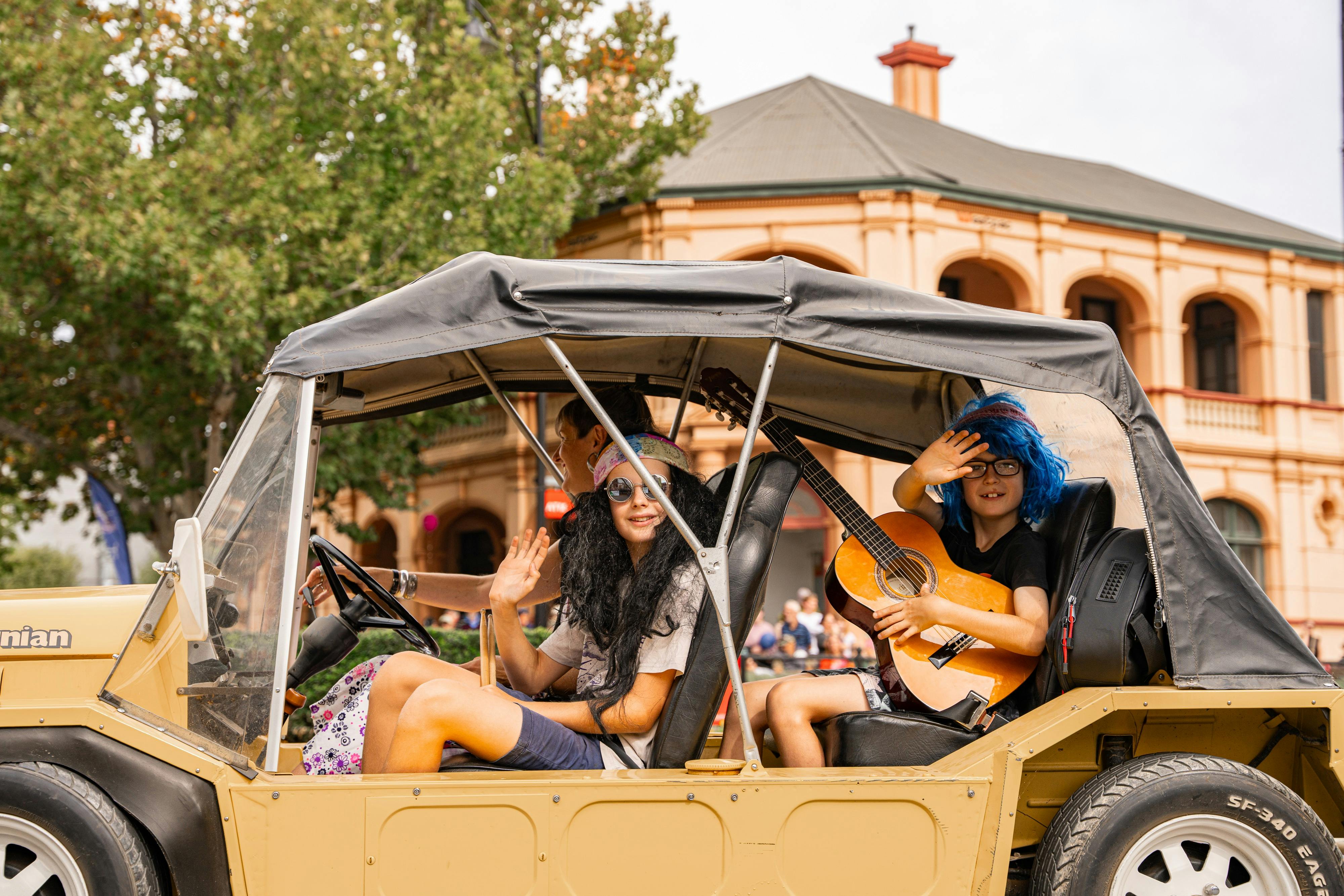 A woman driving a mini moke with kids dressed in 1970s costumes, one has a guitar
