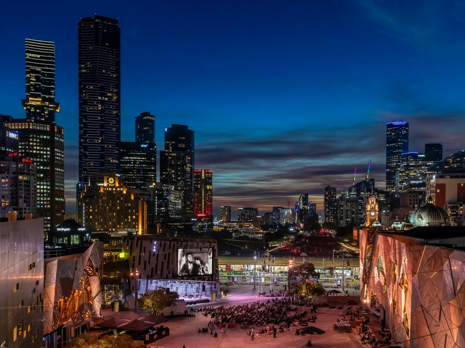 A crowd of people gather in Fed Square at night to watch a film on the big screen