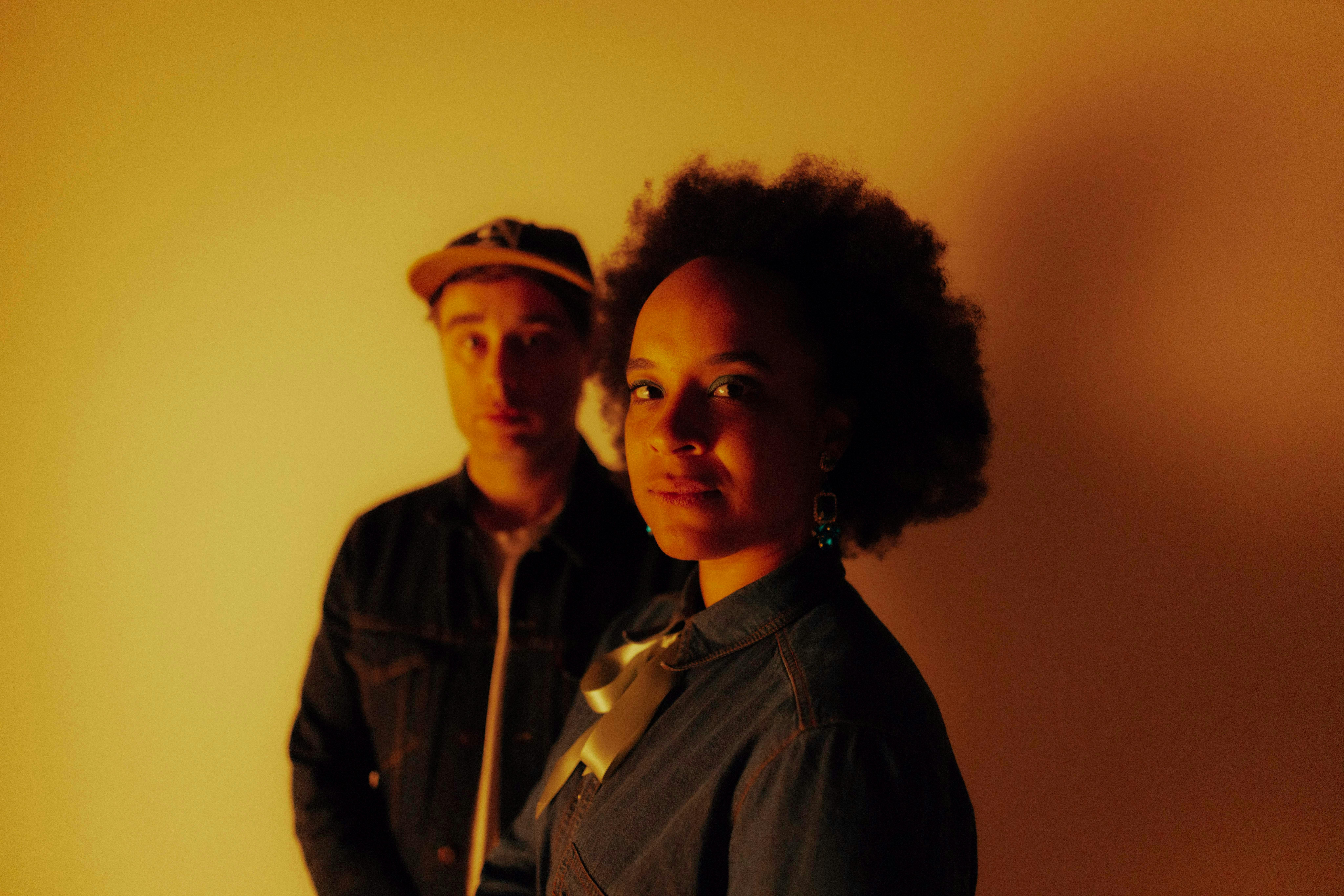 Two musicians in amber light: a woman in denim looks at the camera, a man in a cap behind her.
