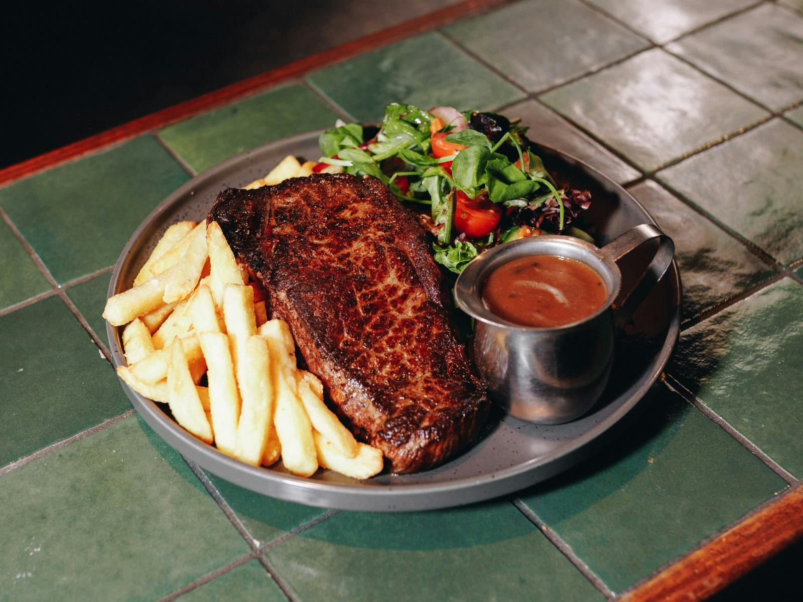 Plate with grilled steak, thick-cut chips, side salad and a small jug of gravy on a tiled bar surfac