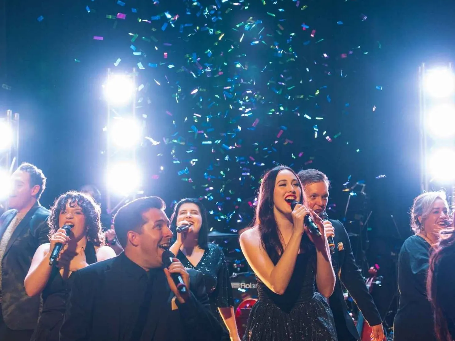 A group of 6 singers with microphones held up to mouth dressed in formal black singing