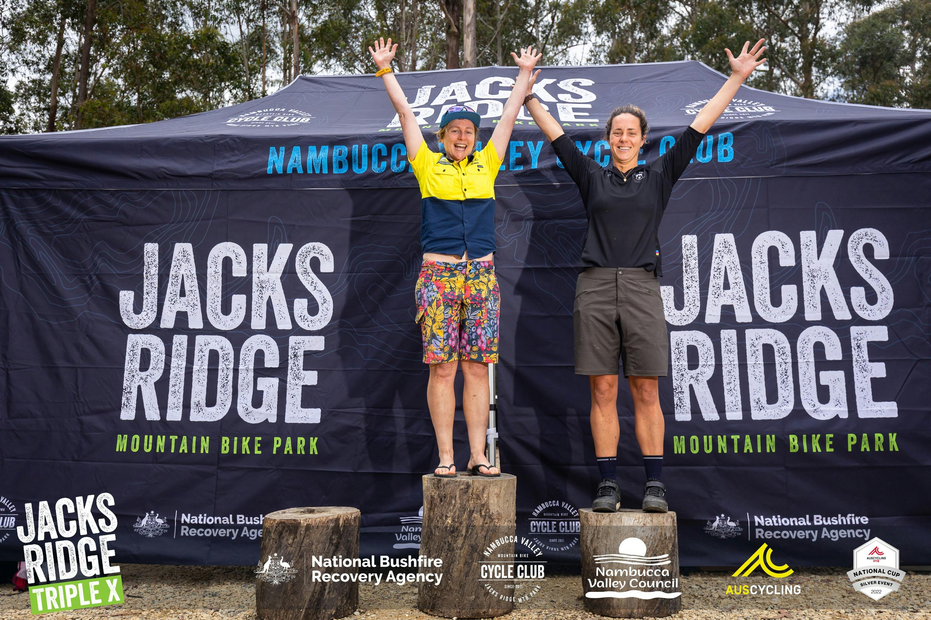 Two ladies on the podium (which are 3 tree trunks) at the XXX, hand in the air in celebration
