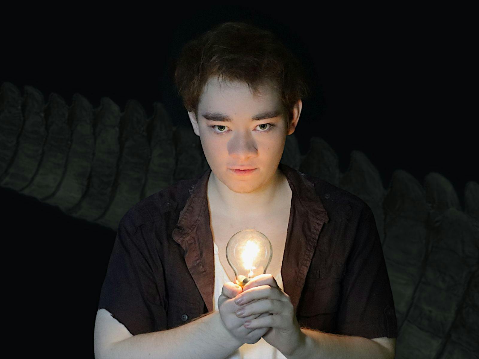 Person dressed as Peter Pan holding a lightbulb,  a crocodile tail in the dark background.