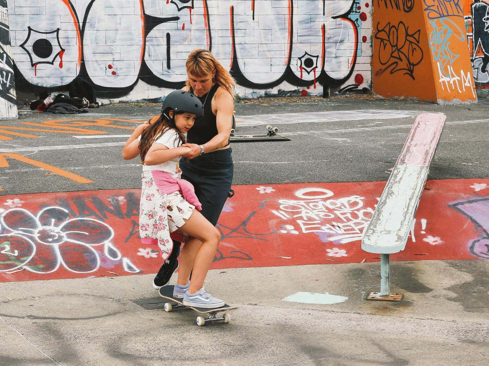 Female presenting skateboard instructor is holding a young girl participant's hands as she rolls