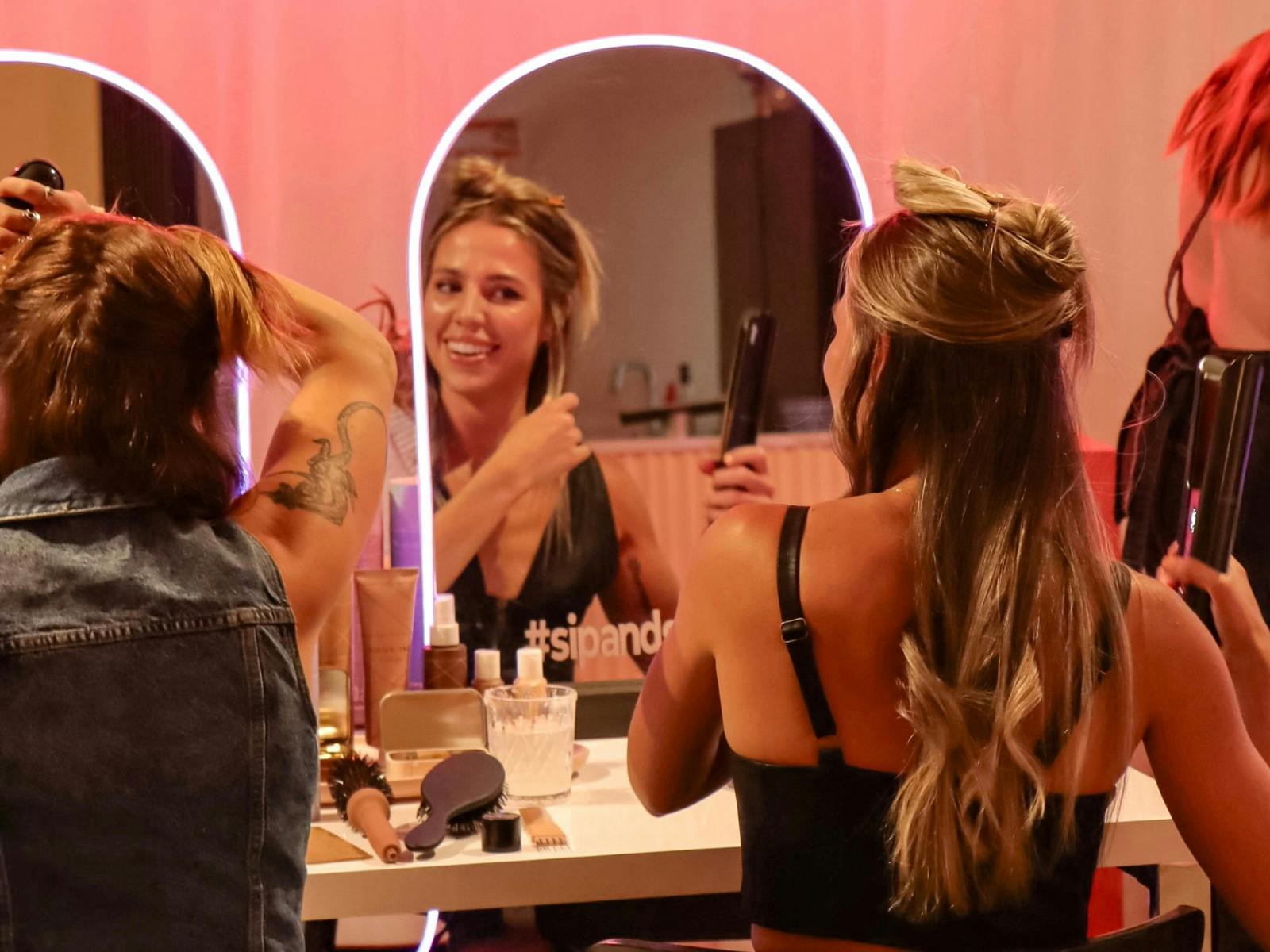 Women styling hair with straighteners at lighted mirrors, smiling in a warm, glowing studio space.