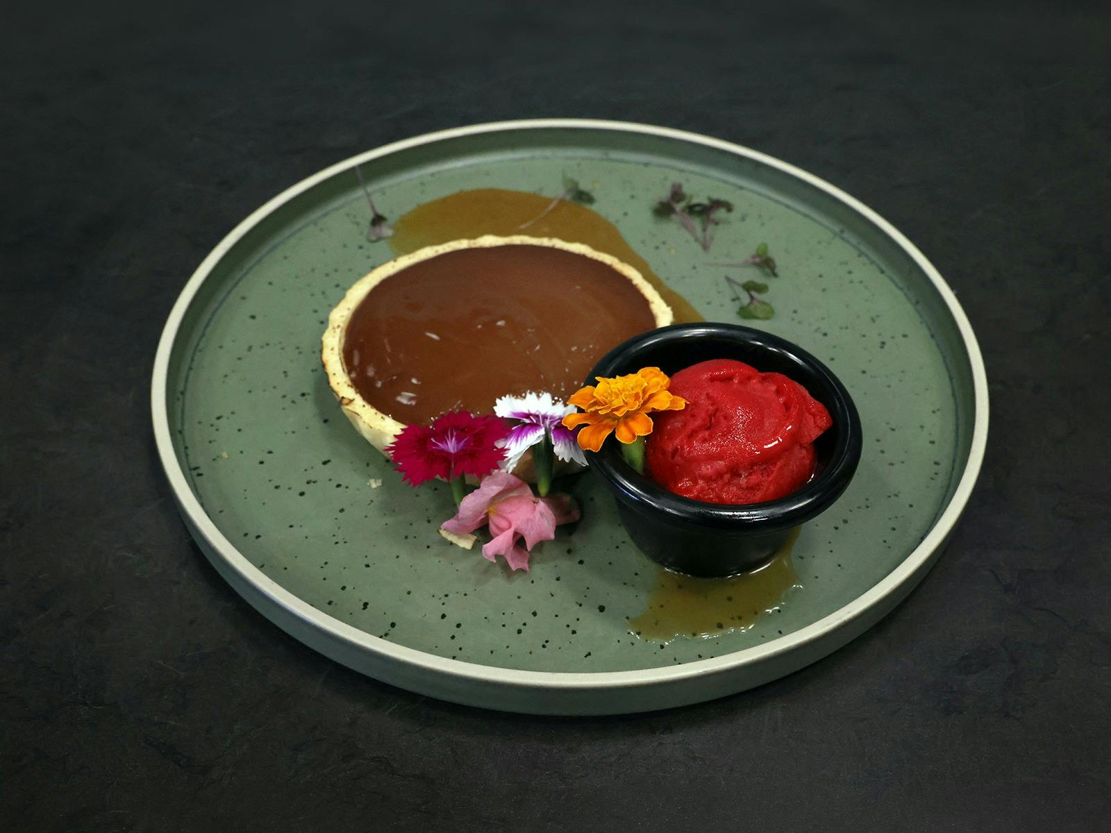 Dark chocolate tart and flowers with raspberry sorbet and salted caramel sauce on a green plate.