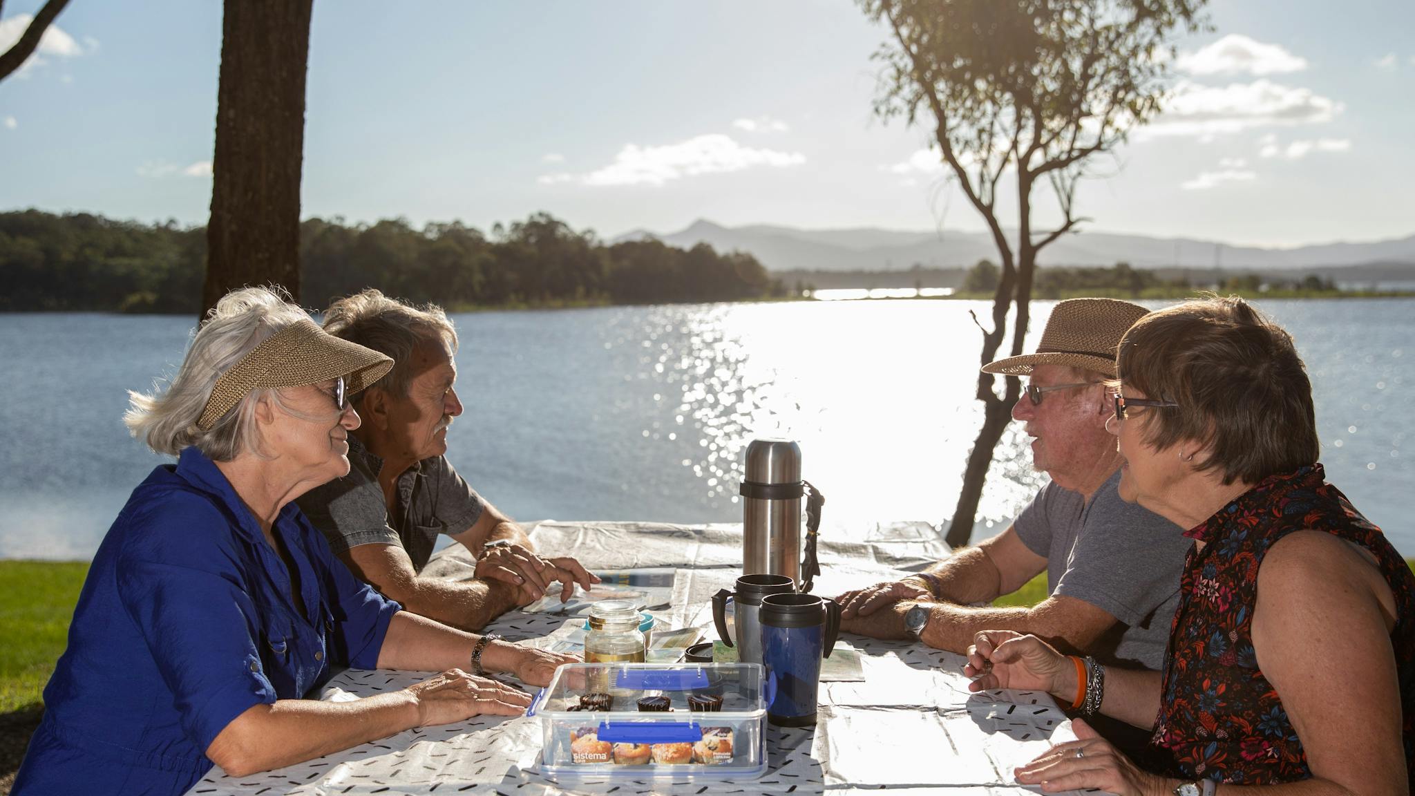 Two elderly couples sitting at a picnic table beside Lake Samsonvale eating and drinking tea