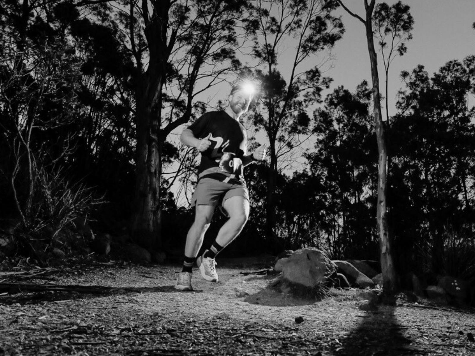 A  black and white image of a male runner at night with a headtorch, passing gum trees