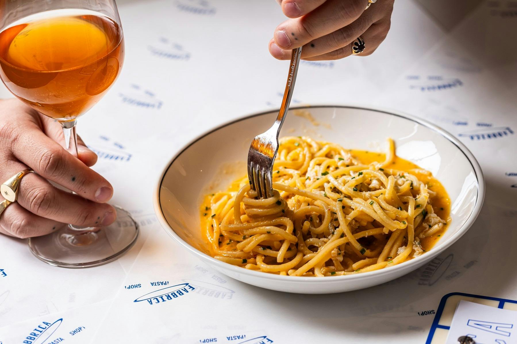 A person holds a glass of orange wine while twirling spaghetti in a white bowl