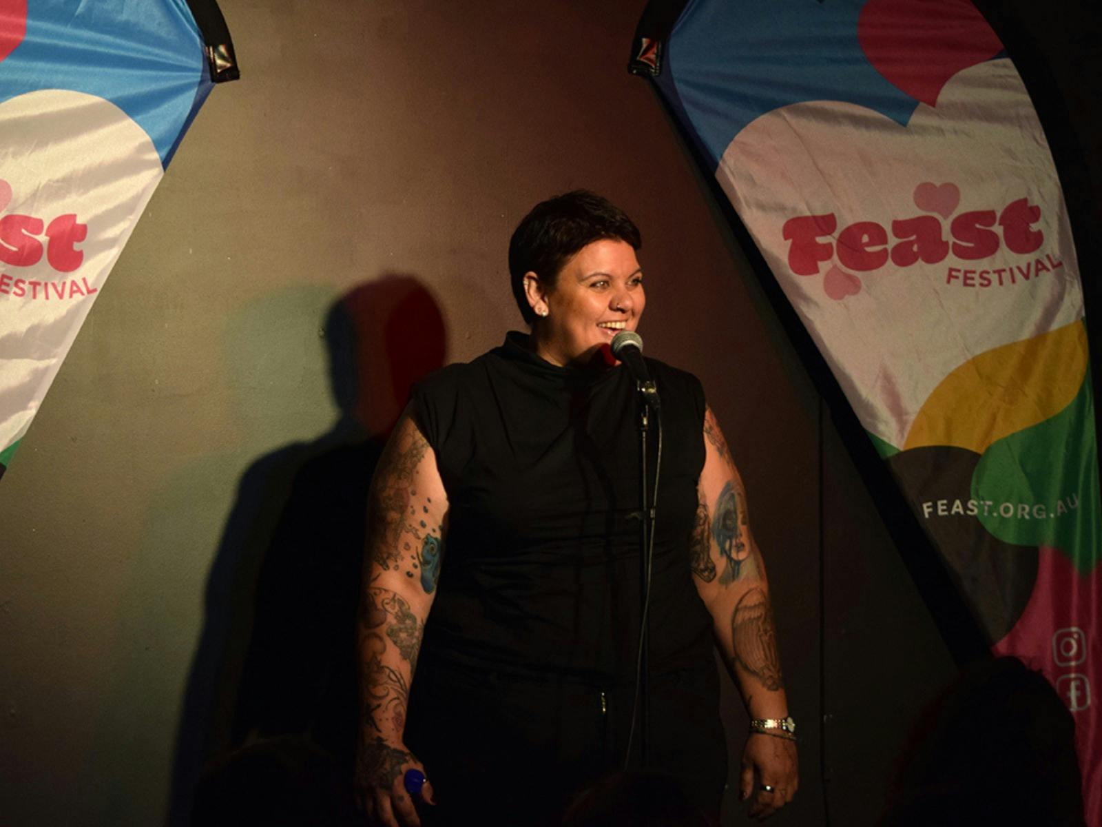 A performer wearing a black sleeveless shirt showing off their tattoos and smiling in front of a mic