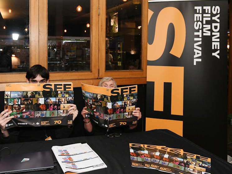 Two people sit at a table holding Sydney Film Festival brochures over their faces