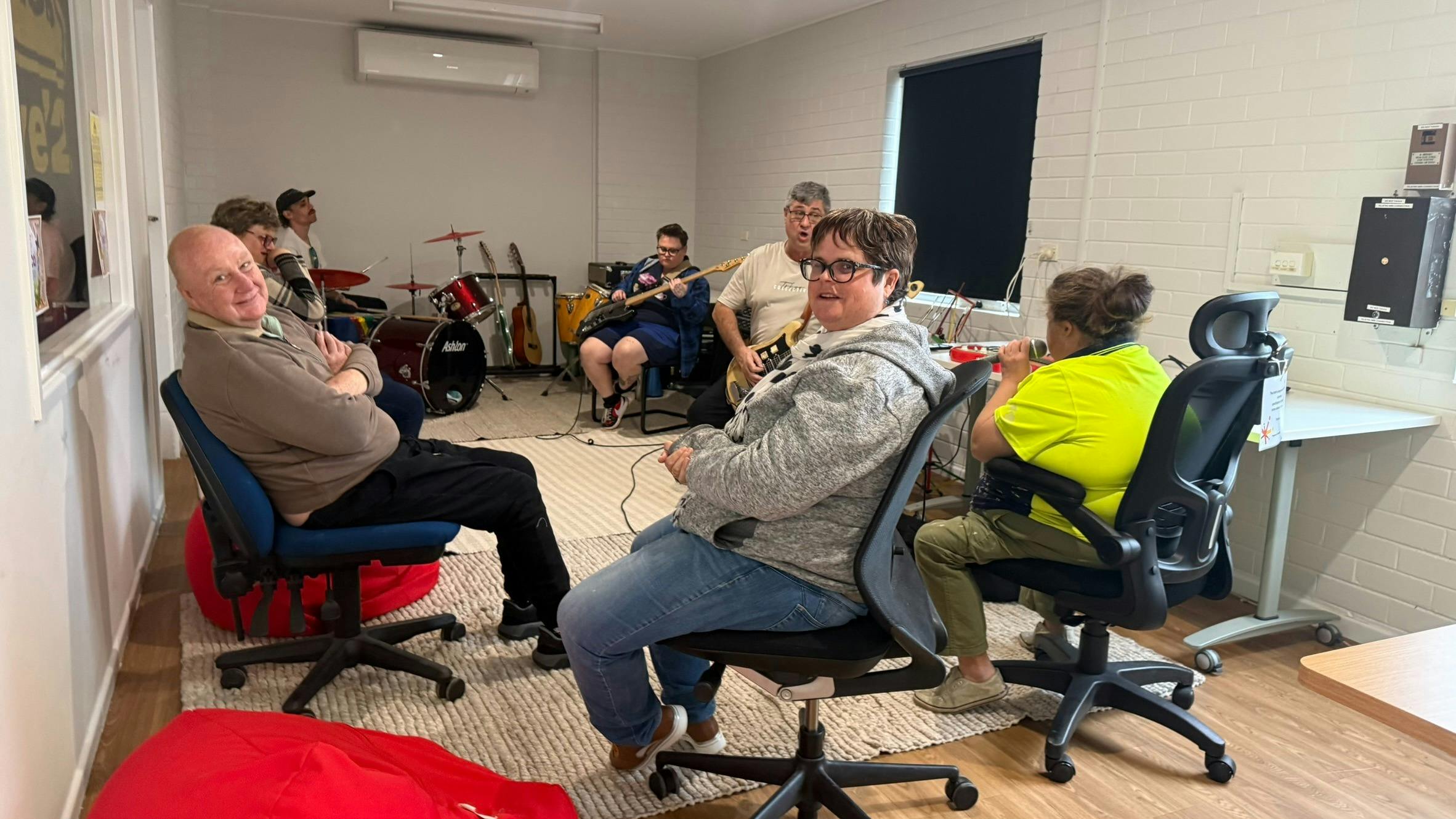 A small group participates in an indoor music session in an office-style room. One person plays an e