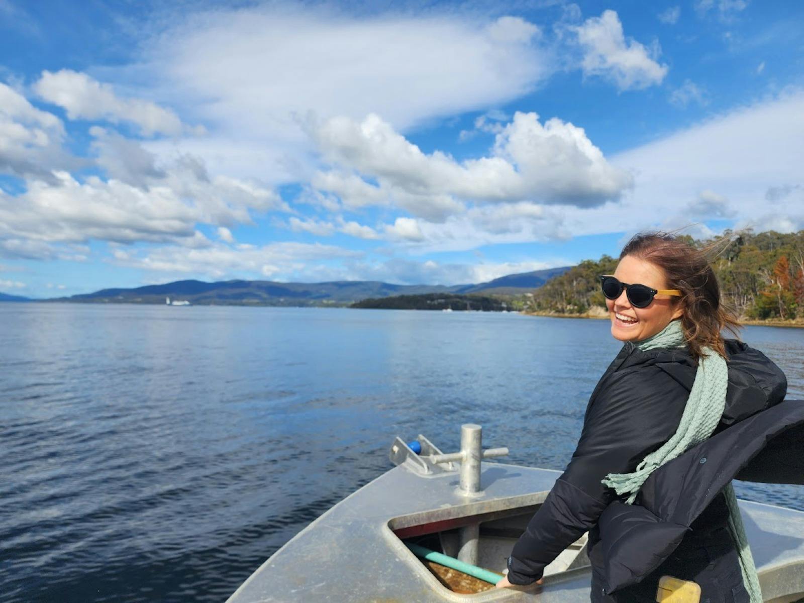 Woman in sunglasses smiles at water, boat railing in foreground and forested shore behind
