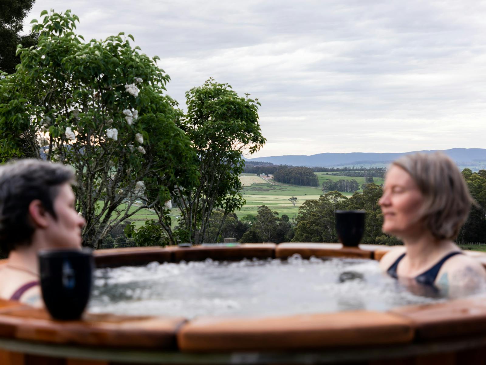 Two women sit talking in a hot tub, in the distance is a beautiful valley and mountains
