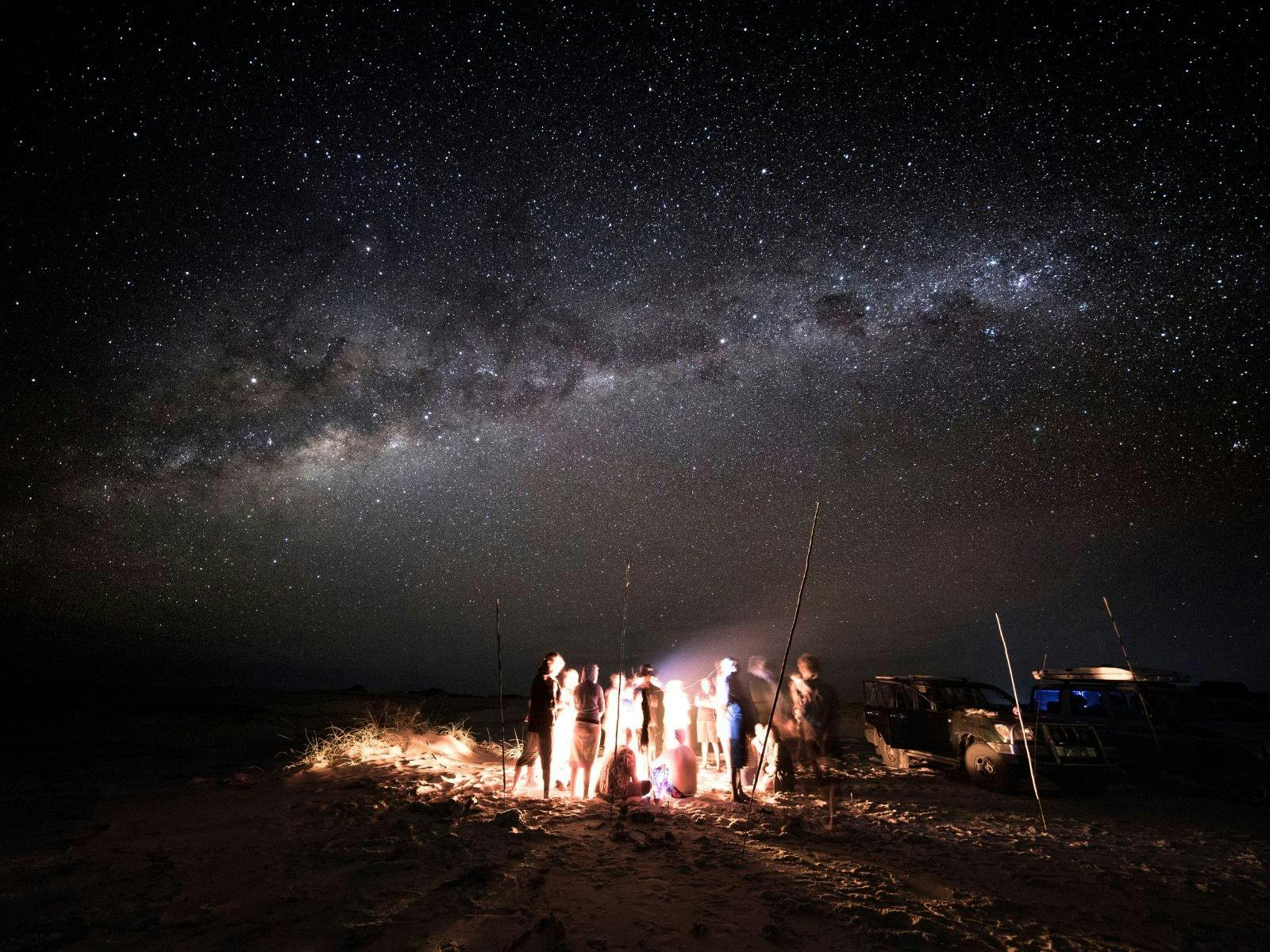 A group of people standing under the milky way around a campfire.