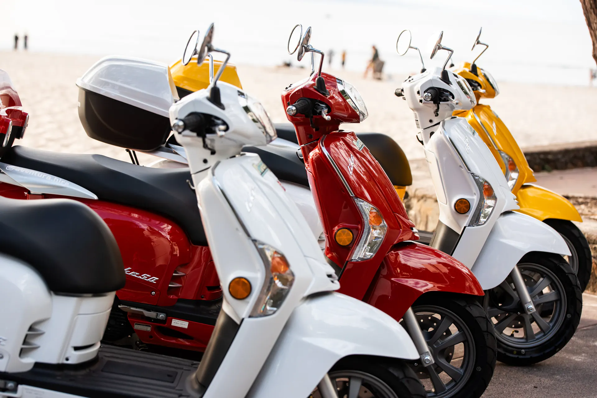 Close up photo of 4 different coloured scooters lined up with beach in background in Noosa