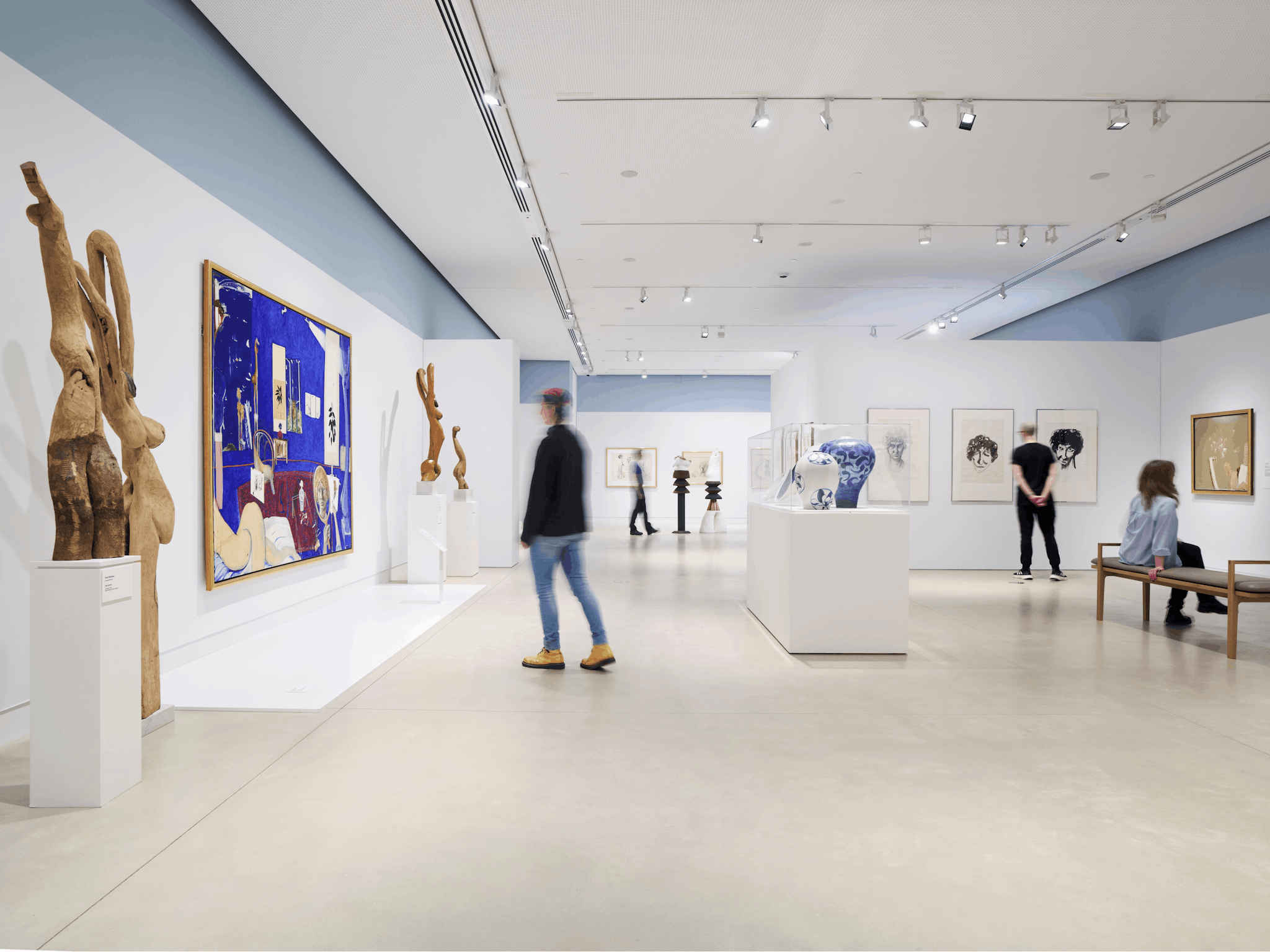 People stand in a large gallery viewing large paintings, wooden statues, and ceramic vessels