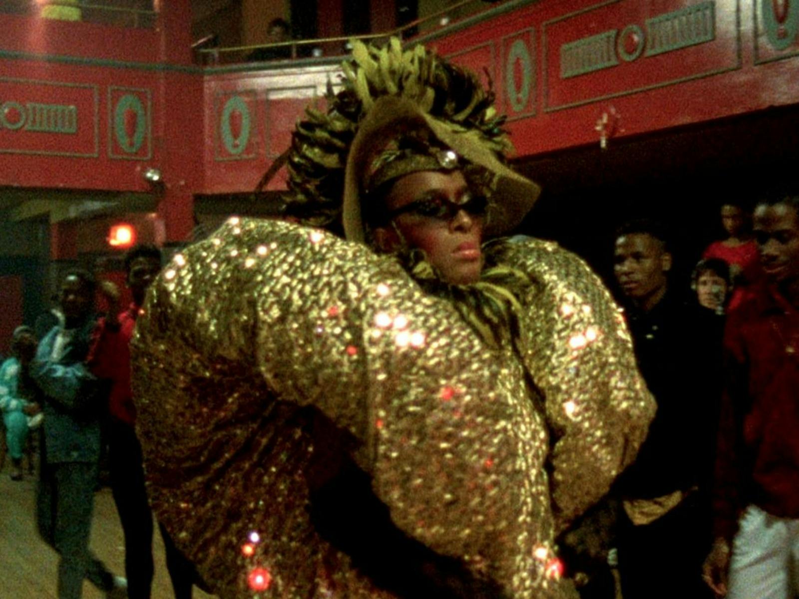 A performer in an extravagant, gold-sequined outfit and headpiece walks in a vibrant ballroom.